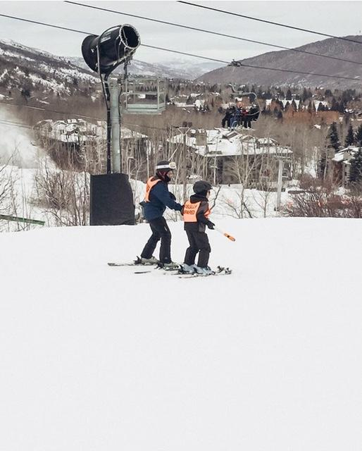 Ashton and his ski guide heading down the mountain. They are wearing bright orange vests. One reads Guide, the other says Visually Impaired.