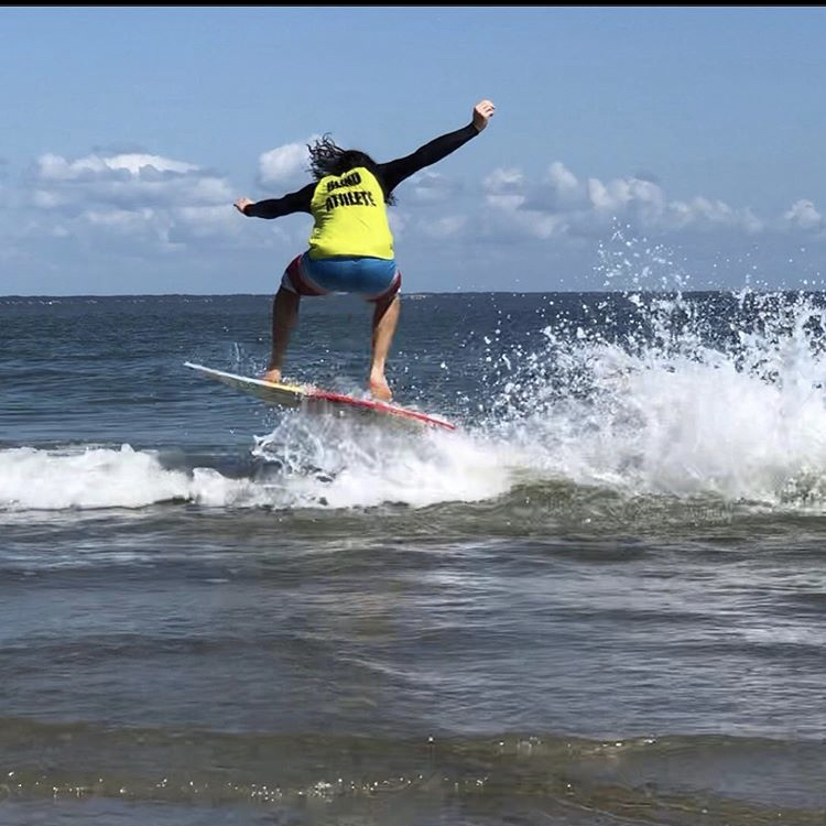 An image of Kai skimboarding and catching air off the waves. He's wearing a jersey which reads "Blind Athlete" across the back.
